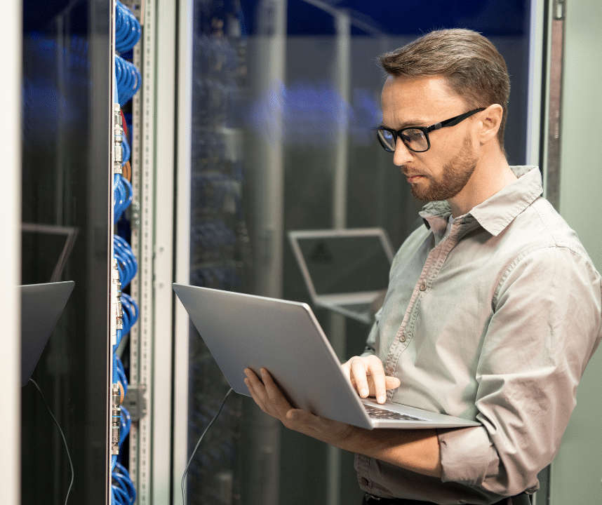 An image of a man working as an Information Technology (IT) specialist standing in a server room. He holds a laptop connected to the servers as he configures and optimizes them.